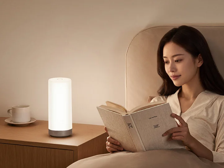 Лампа Mijia Bedside Lamp 3 на прикроватной тумбе, рядом женщина с книгой в постели